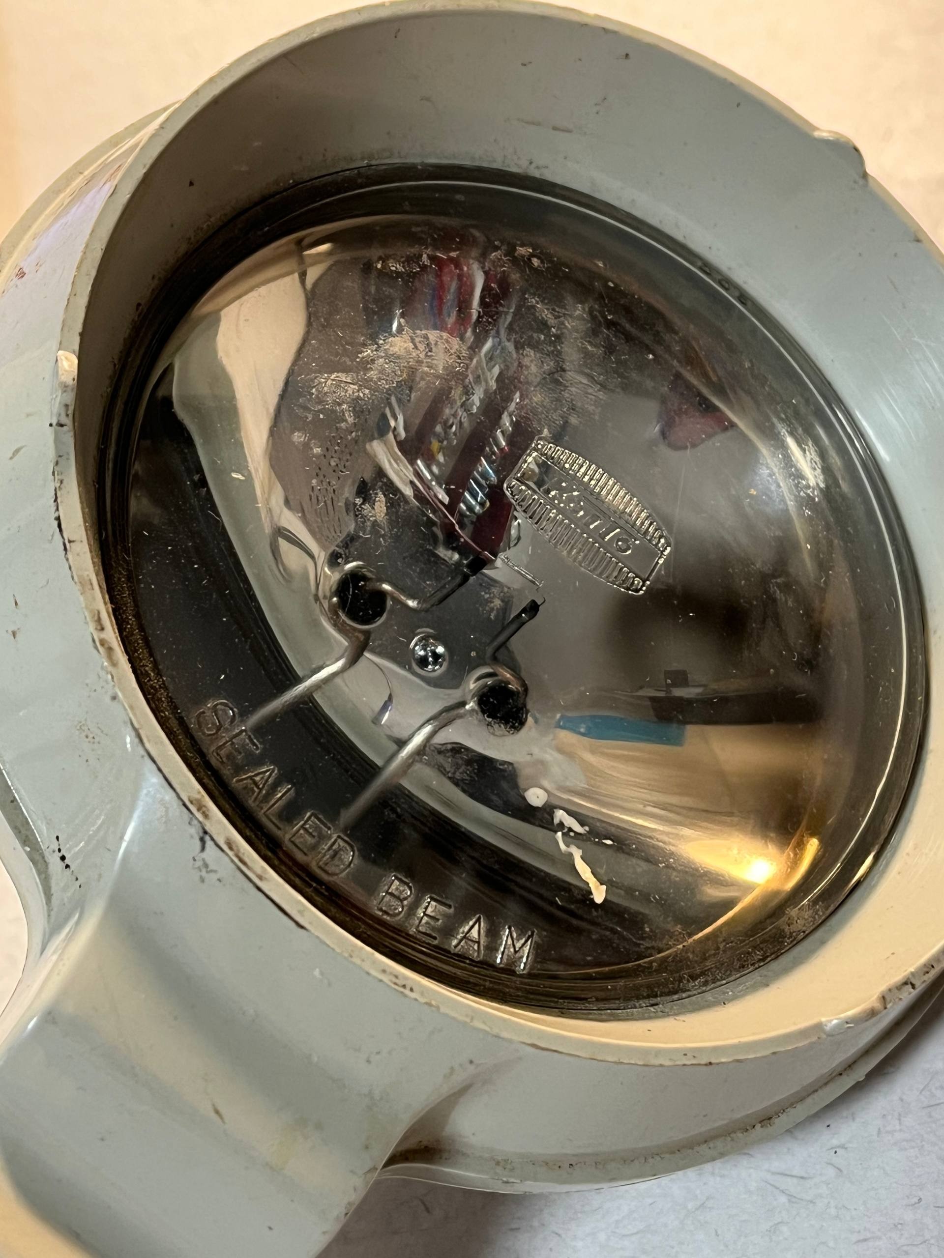 This is a close-up, detailed photograph of a vintage Kaito hand-held torch made in Japan, showing signs of age and use.

The image focuses tightly on the front lens and a portion of the torch's casing. The lens, which appears to be made of glass, is reflective and shows smudges, scratches, and some debris, suggesting it is not new. Inside the lens, metallic components and wires are visible, along with a notable reflection of what might be a red and blue striped object, possibly fabric or paper. There is also a small, rectangular emblem with ridged edges, which is likely the Kaito branding, inscribed within the lens. The words "SEALED BEAM" are clearly embossed around the inner edge of the lens.

The torch casing is a light, possibly off-white or pale beige color, and seems to be made of metal or a durable plastic. The surface of the casing shows some wear, with minor scuffs, a few small dark specks, and what looks like a small chip or dent on the upper right rim of the lens housing. The overall texture of the casing appears slightly matte, with some areas showing a faint sheen. The mounting points for the lens, visible as small metal prongs and a central screw, are also present.