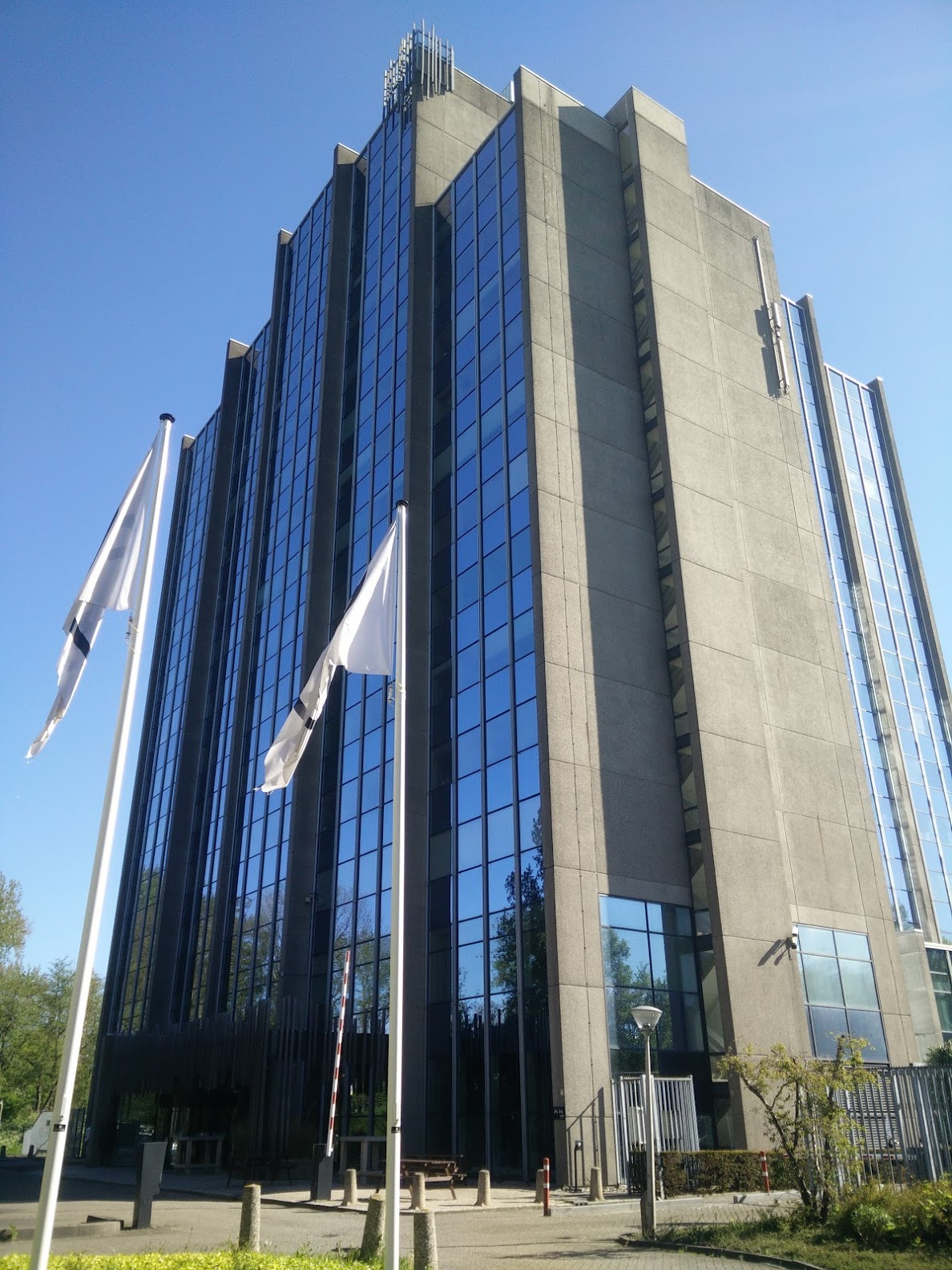 A tall, modern office building with a facade of blue glass windows and concrete walls stands under a clear blue sky. Two white flags on tall poles fly in front of the building.